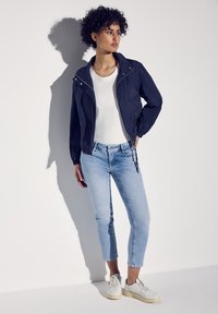 Rompevientos de color navy con cremallera en la parte delantera, dos bolsillos laterales y puños elásticos, combinado con jeans cortos de color azul claro y zapatillas blancas.