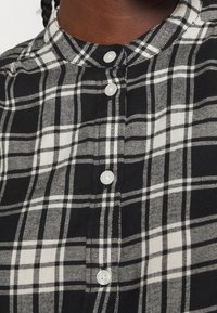 Svart- och vitrutigt knapp-up skjorta med mandarin-krage, som har vita knappar och en mjuk textur. Traditionell rutmönsterdesign.