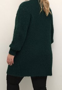 Lang, mørkegrøn strikket cardigan med et tekstureret, ribbet mønster og en let oversized pasform. Har lange ærmer og en høj halsudskæring.
