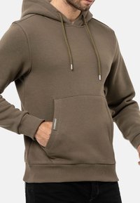 Kapuzensweatshirt in Olivgrün, aus weichem Material gefertigt. Es verfügt über eine Kängurutasche, gerippte Bündchen und einen verstellbaren Kordelzug mit Metallspitzen.