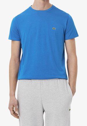 Homme portant un t-shirt bleu à manches courtes et un pantalon de jogging gris clair avec des logos crocodile verts sur la poitrine et la cuisse.
