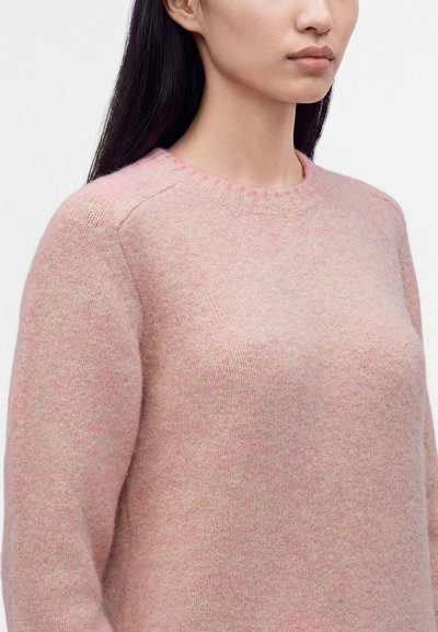 Femme aux longs cheveux noirs portant un pull en tricot doux rose clair avec un col rond sur un fond clair uni.