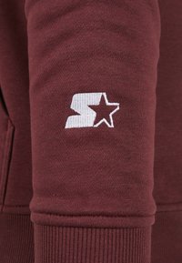 Manica di una felpa color bordeaux con un logo ricamato bianco e una stella. La texture è liscia con polsini a costine.
