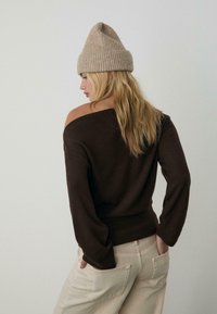 Brauner Off-Shoulder-Pullover mit langen Ärmeln und geripptem Saum, kombiniert mit hellbeigen, hoch taillierten Hosen. Warme beige Strickmütze.