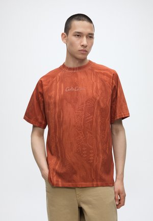Ung mand med kort hår bærer orange tekstureret kortærmet skjorte med "Carlo Colucci" logo og beige bukser, stående med den ene hånd i lommen.