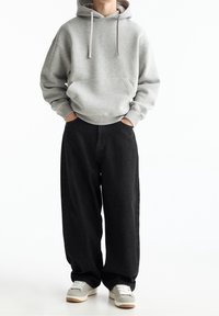 Grijze capuchontrui met voorzak, gecombineerd met loszittende zwarte spijkerbroek en grijze sneakers met witte zolen.