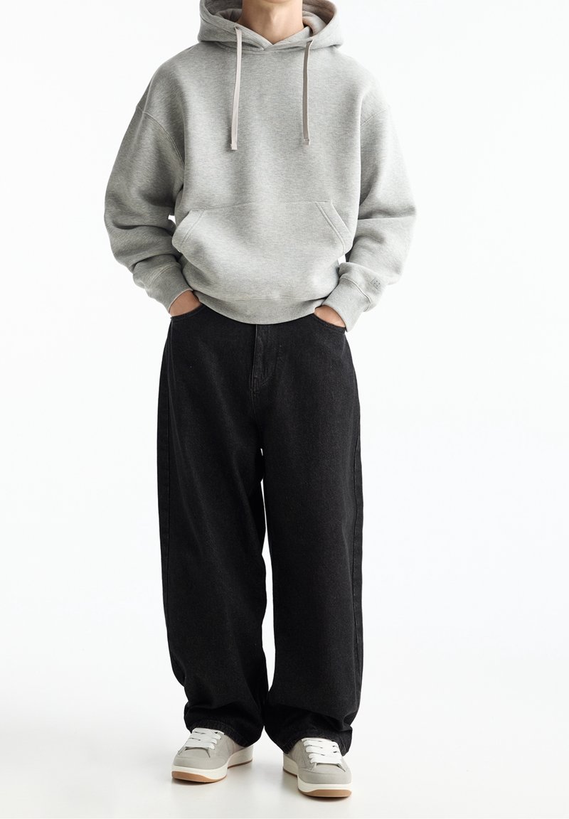 Grijze capuchontrui met voorzak, gecombineerd met loszittende zwarte spijkerbroek en grijze sneakers met witte zolen.