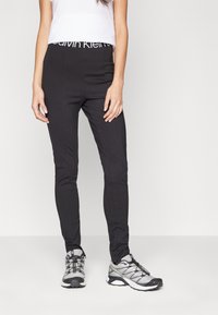 Schwarze, eng anliegende Hose mit einem Calvin Klein Logo-Bund, kombiniert mit grauen Sportschuhen. Die Hose hat eine glatte Textur und ein tailliertes Design.