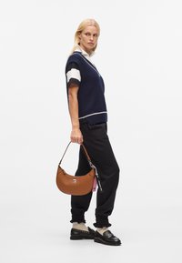 Borsa a tracolla in pelle marrone con una forma curvilinea, accentuata da un logo argentato e un'etichetta rosa. Il modello indossa un gilet di maglia blu navy e pantaloni neri.
