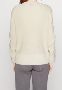 Crèmekleurige gebreide trui met geribbelde textuur, zakken aan de schouders en een brede geribbelde zoom. Getoond vanaf de achterkant, gedragen met grijze geribbelde broek.