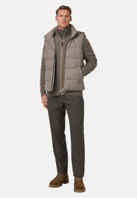 Beige quiltad väst med huva, över en brun polotröja, kombinerad med mörkgrå byxor och bruna mockabutiker. Texturerade tyger synliga.