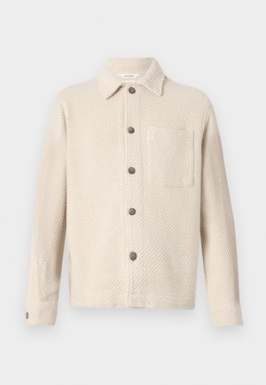 Giacca button-up beige con texture, maniche lunghe, colletto, tasca sul lato destro del petto e bottoni scuri lungo la parte frontale e sui polsi.