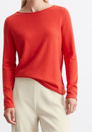 Femme portant un pull ajusté à manches longues de couleur orange vif, associé à un pantalon ample de couleur crème, debout devant un fond uni.