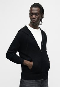 Sweat à capuche noir à fermeture éclair avec cordon de serrage à la capuche, poches avant et poignets côtelés, porté sur un t-shirt blanc, sur un mannequin devant un fond clair.