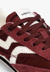 Zapatillas maroon con parte superior de tela y acentos de ante, diseño zigzag blanco y cordones texturizados. La lengüeta muestra la marca "Scalpers Co."