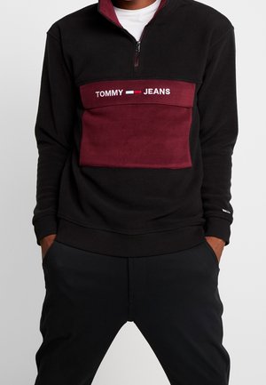 Osoba nosząca czarną i bordową bluzę Tommy Jeans z półzamkiem, z rękami w kieszeniach, na jednolitym jasnym tle.