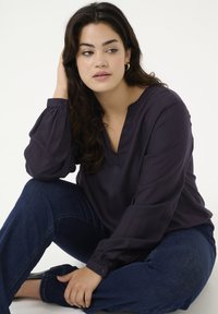 Navy blue blouse in smooth fabric, featuring a v-neck and long, puffed sleeves with buttoned cuffs. Worn with dark denim jeans.