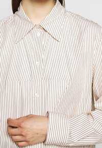 Vaaleanpunainen napillinen paita, jossa on ohuita ruskeita pystyraitoja, terävä kaulus ja pitkät hihat. Paidassa on rintatasku ja napilla kiinnitettävät hihansuut.