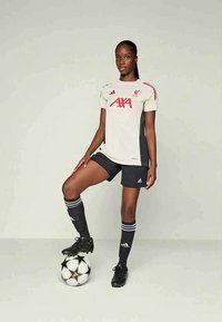 Frau im Liverpool-Fußballtrikot steht mit einem Fuß auf einem schwarz-weißen Fußball vor einem einfarbigen Hintergrund.