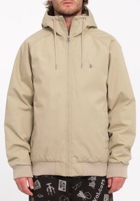 Beige hættetrøje med lynlås foran, snorede detaljer og ribbede håndled. Den har et logo på brystet og en glat, mat overflade.