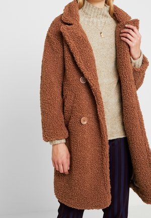 Woman wearing a brown textured teddy coat over a beige turtleneck sweater and dark striped pants, holding the coat's lapel with one hand.