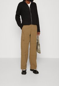 Jersey de punto negro con cremallera, pantalones cargo beige con bolsillos y zapatos gruesos negros. Bolso gris con acentos metálicos. Texturas suaves.