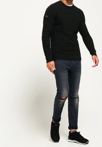 Maglione nero a maglia cable con scollo rotondo, abbinato a jeans scuri strappati e sneakers nere. Presenta una sottile toppa sulla spalla.