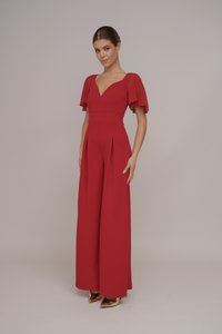 Rode jumpsuit met korte, uitlopende mouwen en een diepe V-hals. Kenmerkt zich door een getailleerde lijfje en brede benen met plooien. Gouden puntige hakken.