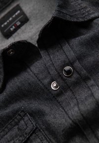 Chemise en denim noir avec une fermeture à boutons, des boutons métalliques et des détails de couture verticale fine. Texture douce et design décontracté.