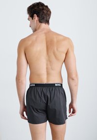 Boxer shorts noirs avec une ceinture élastique arborant la marque "BOSS" en lettres blanches. Le tissu semble lisse et léger.
