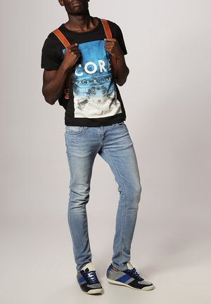 T-shirt noir avec impression graphique bleue, jean bleu clair et baskets avec des accents bleus. Bretelles de sac à dos marron visibles. Tenue décontractée.