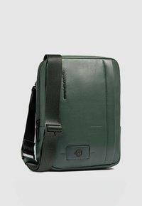 Borsa messenger verde in pelle sintetica con superficie strutturata, tracolla scura regolabile e dettaglio logo frontale. Forma rettangolare con design di cuciture sottili.