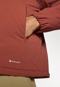 Veste pullover légère rouge avec des poignets élastiqués, une poche latérale zippée et un logo en bas par Snow Peak. Texture lisse.