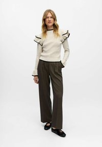 Pull crème côtelé avec des volants bordés de noir ; associé à un pantalon large marron taille haute ; chaussures noires, mains dans les poches.