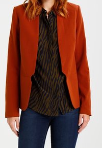 Rostfärgad kavaj med en strukturerad design, slät textur och öppen framsida, som är kombinerad med en mörkgrön randig skjorta.