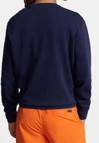 Person som bär en mörk marinblå långärmad skjorta och klara orange byxor med en liten "Polo"-etikett på bakfickan, står med händerna delvis i fickorna.