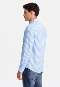 Camisa de manga larga a rayas azul y blanca con cuello abotonado, que presenta un dobladillo curvado y un bolsillo en el pecho, hecha de tela ligera.