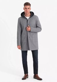 Abrigo texturizado gris con capucha negra, cierre de botones y dos bolsillos frontales; combinado con jeans oscuros y botas de tobillo marrones.