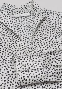Chemise blanche à col avec des taches noires irrégulières, poignets pliés avec deux boutons blancs, et étiquette de marque ETERNA visible.