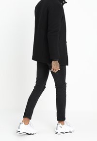 Svart, lång kappa med knappar, bärs över svarta, slitna skinny jeans. Matchas med vita sneakers med grå detaljer.