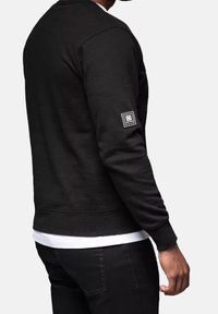 Sudadera negra con un acabado texturizado, puños y dobladillo acanalados, y un parche de logo blanco en la manga izquierda. Lleva una camiseta blanca debajo.