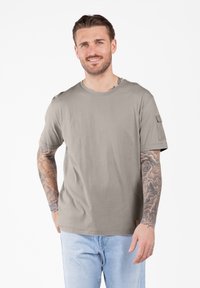 Hombre sonriente con los brazos tatuados, vistiendo una camisa de manga corta color marrón claro y jeans de color azul claro, de pie frente a un fondo blanco.