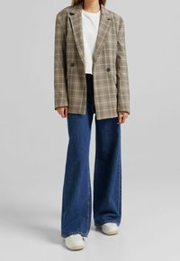 Beżowa marynarka w kratę z podwójnymi guzikami i klapami, noszona na białej bluzce, w zestawieniu z niebieskimi dżinsami o szerokich nogawkach i białymi trampkami.