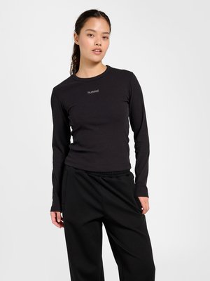 Młoda kobieta ubrana w dopasowaną czarną prążkowaną bluzkę z długim rękawem z logo "hummel" oraz luźne czarne spodnie, stojąca na jednolitym szarym tle.