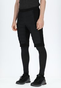 Sorte hybrid shorts lavet af isolerende stof, med en tætsiddende pasform, lynlås-lomme og kombineret med mørke termoleggings og sko.
