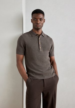 Camisa polo de punto marrón con mangas cortas, que presenta una textura acanalada y una tapeta de botones. Llevada con pantalones marrones, de pie contra un fondo claro.