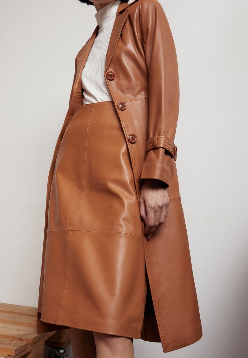 Manteau en cuir fauve à coupe ajustée, avec deux boutons et poignets ceints d'une ceinture, assorti à une jupe en cuir coordonnée. Texture lisse, conception longueur genoux.