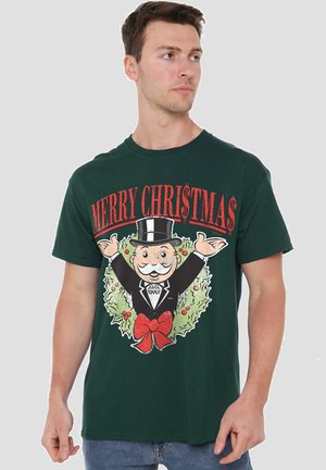 Grünes Baumwoll-T-Shirt mit einem Weihnachtsdesign, das eine Figur in einem schwarzen Smoking zeigt, die die Arme weit ausbreitet und von einem Kranz sowie rotem Text umgeben ist.