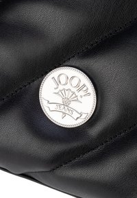 Czarna torba ze strukturalnej skóry z okrągłym srebrnym logo z napisem "JOOP! JEANS" oraz elementami dekoracyjnymi. Miękka, pikowana konstrukcja.