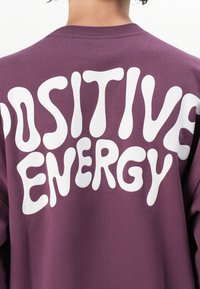 Fialová mikina s veľkým bielym textom v bublinkovom štýle, ktorý na zadnej strane uvádza "POZITÍVNA ENERGIA". Mäkkučký materiál s kulatým výstrihom.